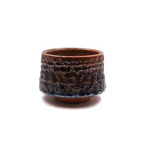 May include: A small, round ceramic bowl with a textured, dark brown and black exterior glaze. The rim and base feature a smooth, reddish-brown finish, while the mid-section displays a bumpy, cratered surface with subtle hints of blue along the bottom edge.