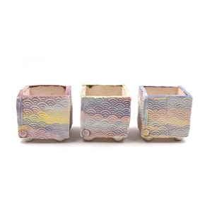 May include: Three square ceramic planters with a wave pattern in shades of pink, yellow, blue, and purple. Each planter has a small circle on the front.