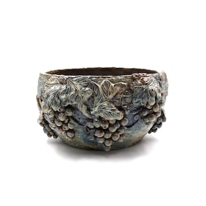 May include: A round ceramic bowl featuring a textured, relief-style design of grape clusters and vine leaves wrapping around the exterior. The surface has a mottled finish with blended tones of deep blue, earthy brown, and muted grey throughout.