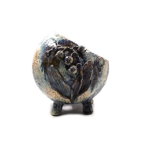 May include: A round, three-legged ceramic planter with a textured, mottled glaze in shades of blue, cream, and dark brown. The exterior features a raised, sculpted relief of lily of the valley flowers and leaves, with a slanted, open top rim.