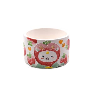 May include: A round, white ceramic planter with a whimsical strawberry design. The planter features a cartoon strawberry character with a winking face, surrounded by red strawberries and green leaves. The planter is suitable for small plants or succulents.