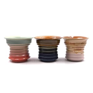 May include: Three ceramic cups with a flared rim and a ribbed, tapered body. The left cup features a light green top fading into a reddish-orange base. The center cup is brown at the top, transitioning to dark blue. The right cup is tan and pink.