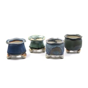 May include: Four small, handcrafted ceramic pots in varying shades of blue and green. Each pot has a unique shape and sits on three small, rounded feet. The pots have a textured, glazed finish, perfect for small plants or succulents.