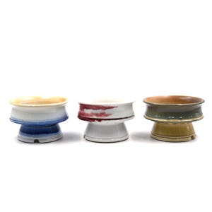 May include: Three ceramic bowls with a pedestal base. The bowls have a wide rim and are painted in shades of blue, white, red, green, and brown. The bowls are displayed on a white background.