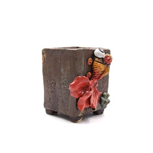 Pode incluir: Um vaso quadrado de cer&acirc;mica castanha com acabamento texturizado. Um design detalhado de peixe dourado tridimensional em laranja, vermelho e branco decora a lateral. O vaso tem p&eacute;s pequenos e &eacute; adequado para plantas pequenas.