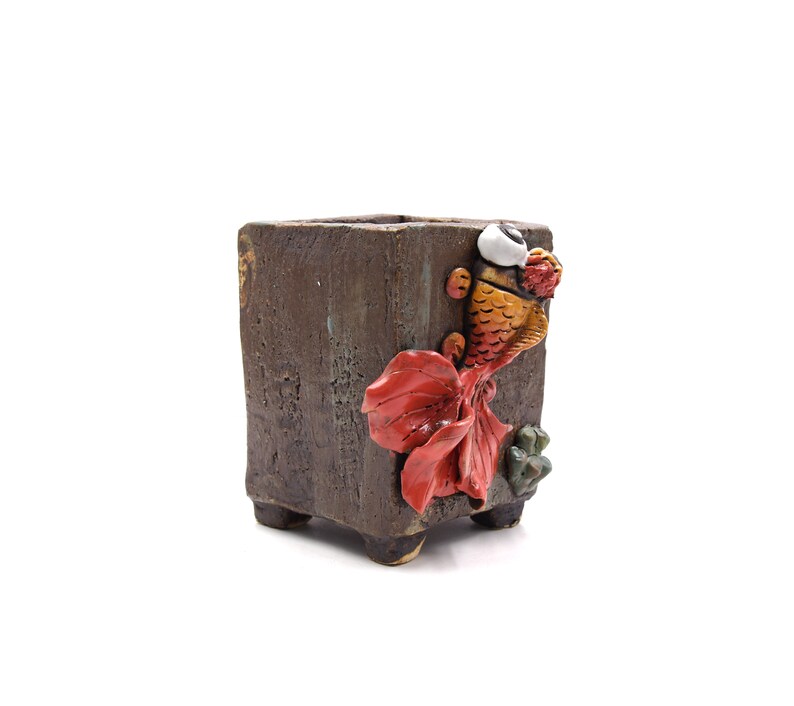 Pode incluir: Um vaso quadrado de cer&acirc;mica castanha com acabamento texturizado. Um design detalhado de peixe dourado tridimensional em laranja, vermelho e branco decora a lateral. O vaso tem p&eacute;s pequenos e &eacute; adequado para plantas pequenas.