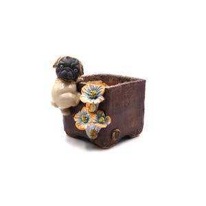 Puede incluir: Una maceta cuadrada de cerámica marrón con una figura de perro pug en el lateral. La maceta tiene detalles de flores azules y amarillas. El pug tiene la cara negra y el cuerpo beige. La maceta está vacía.