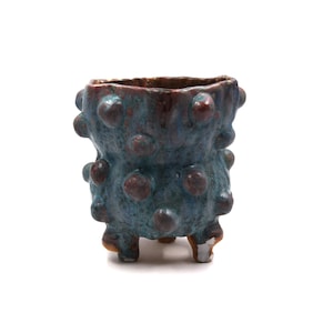 May include: A small, tripod ceramic planter featuring a textured surface with raised, rounded bumps. The vessel is finished in a mottled glaze of deep blue, teal, and earthy brown tones, with a wide, open top and three short, sturdy legs at the base.