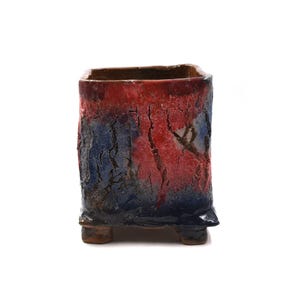 Puede incluir: Una maceta rectangular de cerámica con una superficie texturizada. La maceta presenta un degradado de colores, que va del rojo en la parte superior al azul en el medio y al azul oscuro en la parte inferior. Tiene pequeños pies.