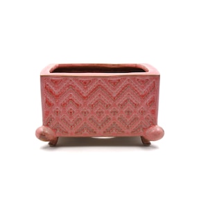 May include: A red ceramic planter, rectangular in shape, with a detailed geometric design. The planter has three small, rounded feet and a textured surface. The interior is a darker shade of red, with a capacity for plants.