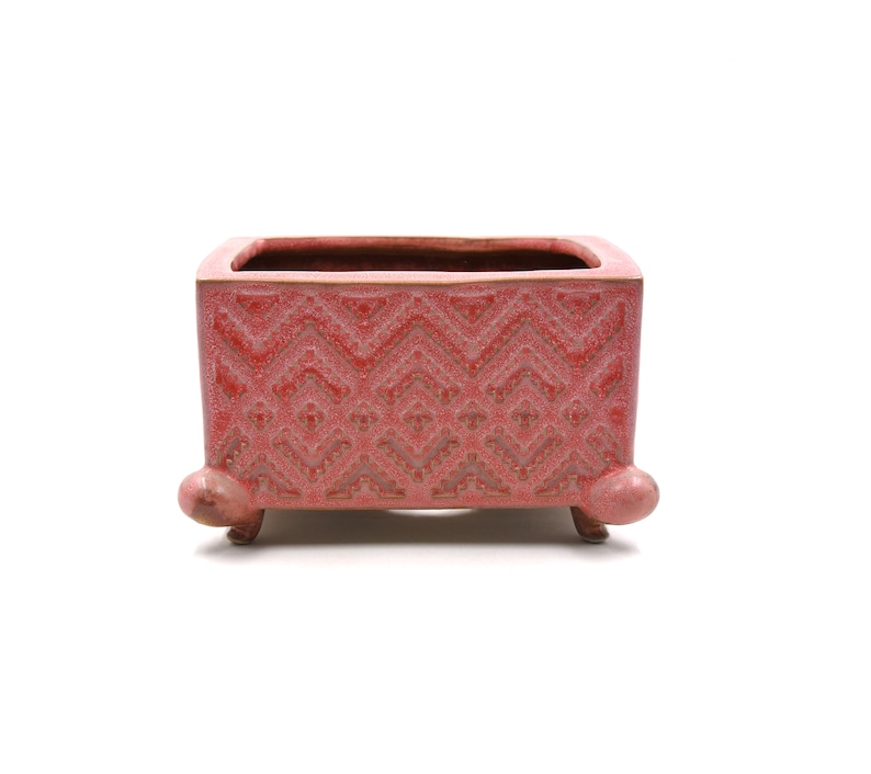 May include: A red ceramic planter, rectangular in shape, with a detailed geometric design. The planter has three small, rounded feet and a textured surface. The interior is a darker shade of red, with a capacity for plants.