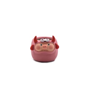 May include: A small, pink ceramic planter shaped like a cartoon devil. The planter has red horns, ears, and a face with googly eyes and a pig-like snout. The planter is designed to hold small plants or succulents.