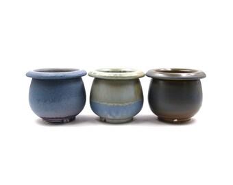 May include: Three ceramic pots in different colors and glazes. The pots are round with a wide opening and a slightly flared rim. The pot on the left is a light blue with a purple hue, the middle pot is a light green with a blue hue, and the pot on the right is a dark green with a brown hue.