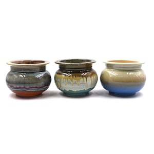 May include: Three small ceramic pots with wide rims and rounded bodies. The left pot features a grey, blue, and orange glaze. The center pot has a brown, cream, and teal drip glaze. The right pot displays a smooth cream and blue gradient finish.