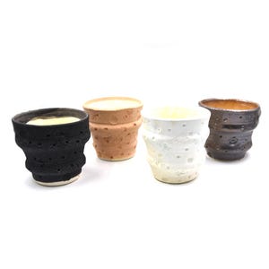 May include: Four handmade ceramic cups in varying colors: black, tan, white, and dark brown. Each cup has a unique textured surface with small, irregular holes. The cups are displayed against a white background.