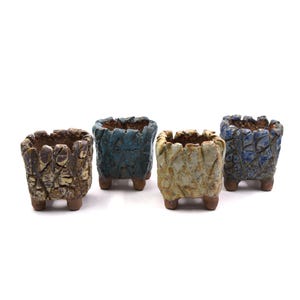 May include: Four small, square ceramic planters in shades of brown, blue, and green. Each planter has a textured, mosaic-like surface and sits on three small feet. The planters are empty and ready for plants.