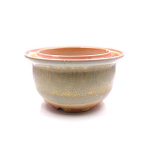May include: A ceramic planter with a light brown glaze and a light pink rim. The planter has a textured surface and a drainage hole.