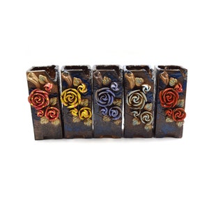 May include: Five small ceramic planters with different colored roses. Each planter is brown with a dark blue glaze and a white background. The roses are red, yellow, blue, beige, and red.
