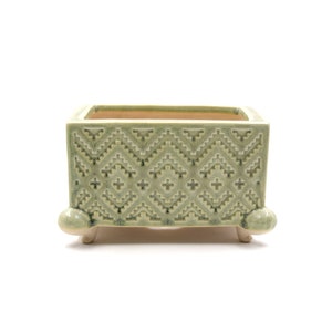 May include: A square, pale green ceramic planter with a geometric design. The planter has a rectangular shape and is supported by four small, rounded feet. The interior is a contrasting tan colour.
