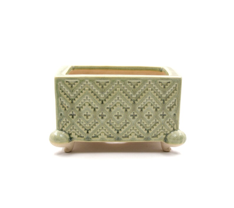 May include: A square, pale green ceramic planter with a geometric design. The planter has a rectangular shape and is supported by four small, rounded feet. The interior is a contrasting tan colour.