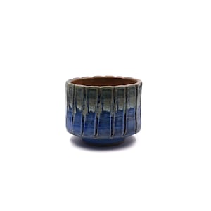 May include: A cylindrical ceramic cup featuring a fluted, vertical ribbed texture. The exterior glaze transitions from a deep, vibrant blue at the base to a muted, earthy grey-green at the rim, with a smooth, unglazed brown interior surface.