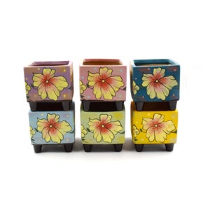 Peut inclure: Six petits pots carrés en céramique avec des motifs floraux. Les pots sont empilés en deux rangées de trois. La rangée du haut présente un pot rose, bleu et jaune. La rangée du bas présente un pot bleu clair, jaune et vert. Chaque pot a un motif floral différent avec des fleurs jaunes.