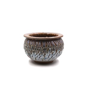 May include: A small, round ceramic bowl featuring a textured, crackled glaze finish. The exterior displays a gradient of colors transitioning from a deep, dark brown at the rim to a lighter, pale blue and white pattern across the body of the vessel.