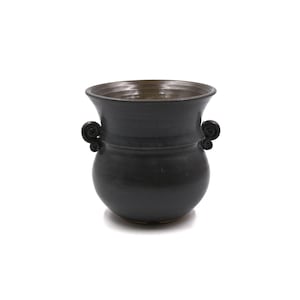 May include: A dark charcoal-colored ceramic pot with a rounded body and a wide, flared rim. The vessel features two small, symmetrical scroll-shaped handles on opposite sides. The interior of the pot has a smooth, dark brown glazed finish.