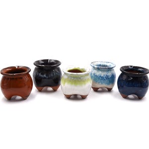 May include: Five small, round ceramic pots in varying colors: brown, black, white with green, blue with white, and dark blue. Each pot has a wide opening and three small feet.