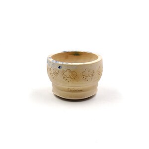 Puede incluir: Un pequeño huevo de cerámica blanca con un diseño floral en relieve. La taza tiene un esmalte azul en el borde y un sello azul en la parte inferior que dice "The Garden".