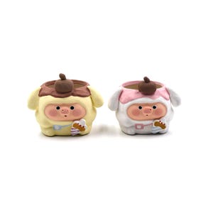 May include: Two ceramic planters in the shape of cartoon characters. One is yellow with a brown top and the other is white with a pink top. Both have a round face with rosy cheeks, a small nose, and a small mouth. Each holds a small dessert.