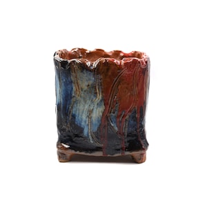 May include: A handcrafted ceramic planter with a textured finish. The rectangular planter has a wavy rim and small feet. It features a colour scheme of blue, brown, and red, with a dripped glaze effect. Measurements are approximately 15 cm high.