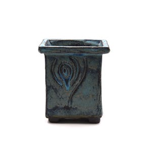 Puede incluir: Una maceta cuadrada de cerámica de color verde azulado, con una superficie texturizada y un diseño decorativo en forma de ojo estilizado en la parte delantera. La maceta tiene un borde oscuro y se apoya en cuatro pequeños pies.