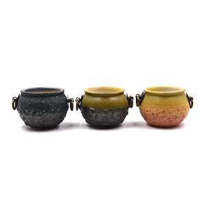 May include: Three small ceramic pots with rounded bodies and wide openings. Each pot features two side handles with attached metal rings. The first is dark gray, the second is green and gray, and the third is yellow and orange with a textured finish.