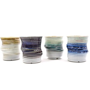 May include: Four ceramic cups with a wavy design and different colored glazes. The cups are arranged in a row from left to right. The first cup is brown and white, the second is purple and white, the third is green and white, and the fourth is blue and white.