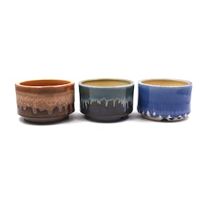 May include: Three cylindrical ceramic pots arranged in a row. The first pot is brown with a tan drip glaze. The second pot is dark blue and green with a white drip glaze. The third pot is bright blue with a white drip glaze at the base.