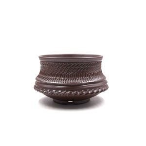 Può includere: Un vaso di terracotta marrone scuro con un motivo decorativo intrecciato attorno al bordo.