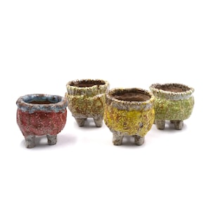 May include: Four small, textured ceramic pots in shades of red, yellow, and green. Each pot has a unique color and texture, with a rough, aged appearance. The pots are elevated on three small legs, suitable for small plants or succulents.