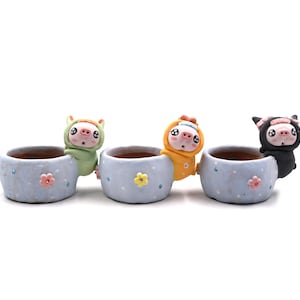 May include: Three light blue ceramic planters with cartoon pig characters in green, yellow, and black outfits. Each planter has a flower design and is suitable for small plants or succulents.
