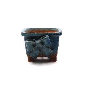 May include: A square, blue ceramic planter with a bow design on the front. The planter has a textured finish and sits on four small feet. The interior is a contrasting brown color.