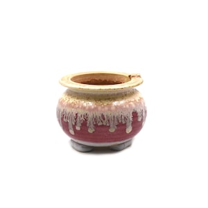 May include: A small, round ceramic pot featuring a wide, flat rim and three short, stubby feet. The exterior displays a deep magenta base color with a thick, cream-colored glaze dripping downward from the top rim, creating a textured, layered effect.