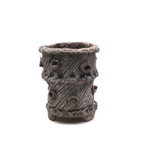 May include: A small, textured, dark gray ceramic planter with a rustic, tree-trunk-like design. The planter has an oval opening and is adorned with carved details, including circular accents and vertical lines, and sits on small feet.