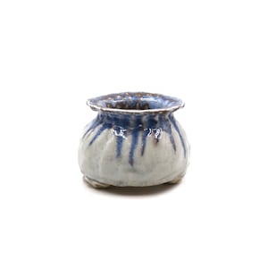 May include: A small, round ceramic planter with a wide, ruffled rim. The exterior features a textured, off-white base glaze with vertical blue drips running from the top rim down the sides. The interior is glazed in a deep, dark blue tone.