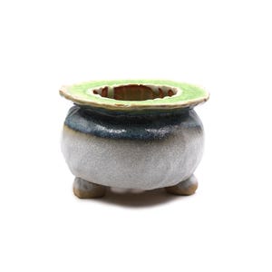 Peut inclure: Un petit pot en céramique rond doté de trois pieds courts. L'extérieur présente une base texturée gris clair qui passe à un vernis bleu foncé près du bord supérieur. Le rebord large et plat est fini avec un vernis vert citron texturé.