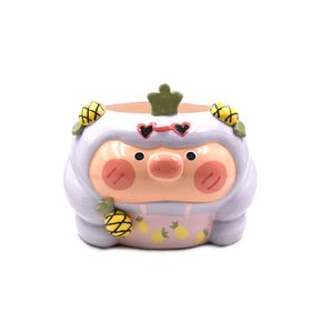 Può includere: Un vaso in ceramica a forma di maialino che indossa un vestito con cappuccio color viola chiaro e occhiali da sole a forma di cuore. Il maialino ha il viso color pesca con guance rosa e piccoli motivi a forma di ananas gialla sul cappuccio.