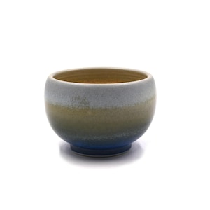 May include: A ceramic bowl with a rounded shape and a gradient color scheme. The bowl transitions from a blue base to a tan and gray upper section, with a light brown interior. The bowl is likely handmade.