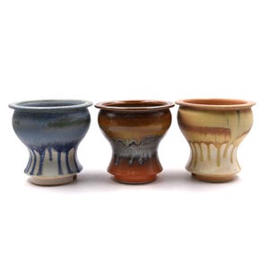 May include: Three ceramic cups with flared rims and tapered bases. The left cup is blue with white drips, the center cup is brown and grey with a glossy finish, and the right cup is cream and tan with vertical glaze drips along the exterior surface.