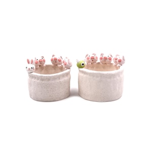 May include: Two cylindrical ceramic planters with a speckled off-white finish. The left planter features a white rabbit and three pink pig figurines on the rim. The right planter features a green creature and three pink pig figurines on the rim.
