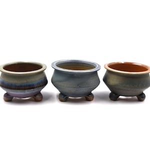 May include: Three small, ceramic bonsai pots with a rounded shape and three small feet. The pots have a glazed finish in shades of blue, green, and brown. The interior of each pot is a contrasting color, with a light brown rim.