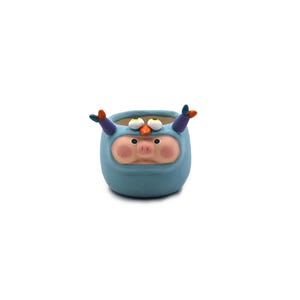 May include: A whimsical, hand-painted ceramic planter in the shape of a cartoon character. The planter is primarily blue with a pig-like face, large eyes, and purple and orange accents. The small pot is designed for small plants.